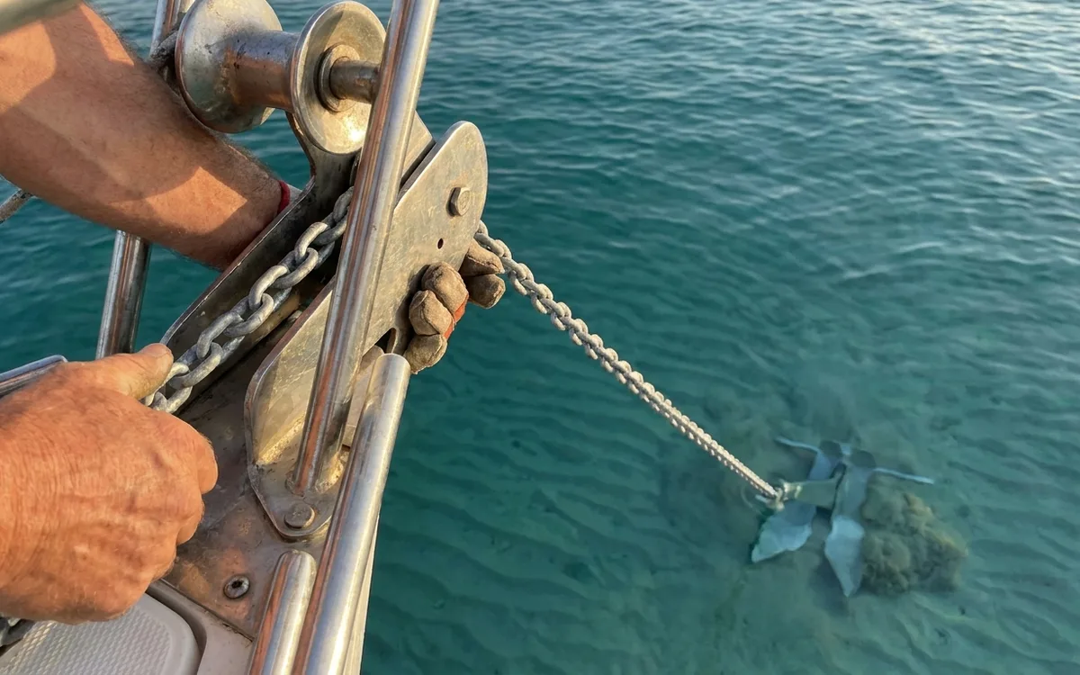 Hands pay out chain over the bow roller as a fluke anchor digs into sandy seabed at a shallow angle.