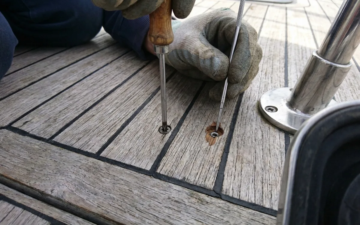 Hands checking an exposed teak deck screw beside a stanchion base for remaining wood thickness.