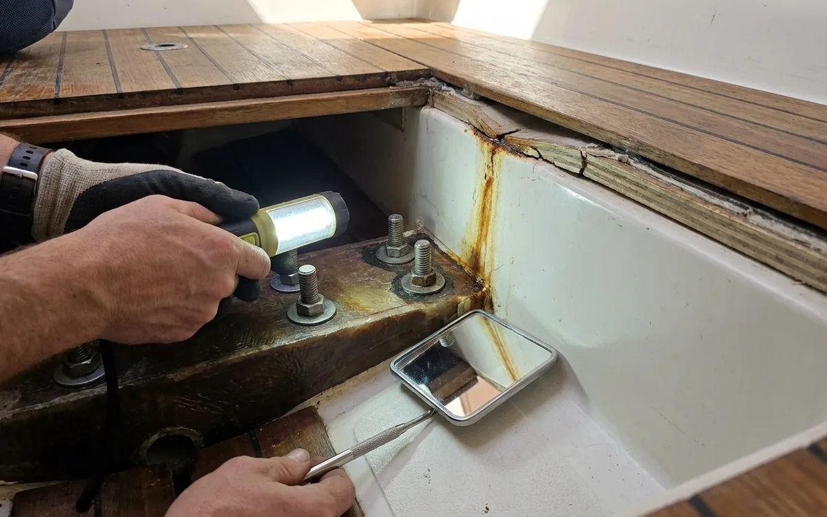 Hands use a flashlight and inspection mirror to check keel bolts and hull-grid bonds beneath the lifted sole boards.
