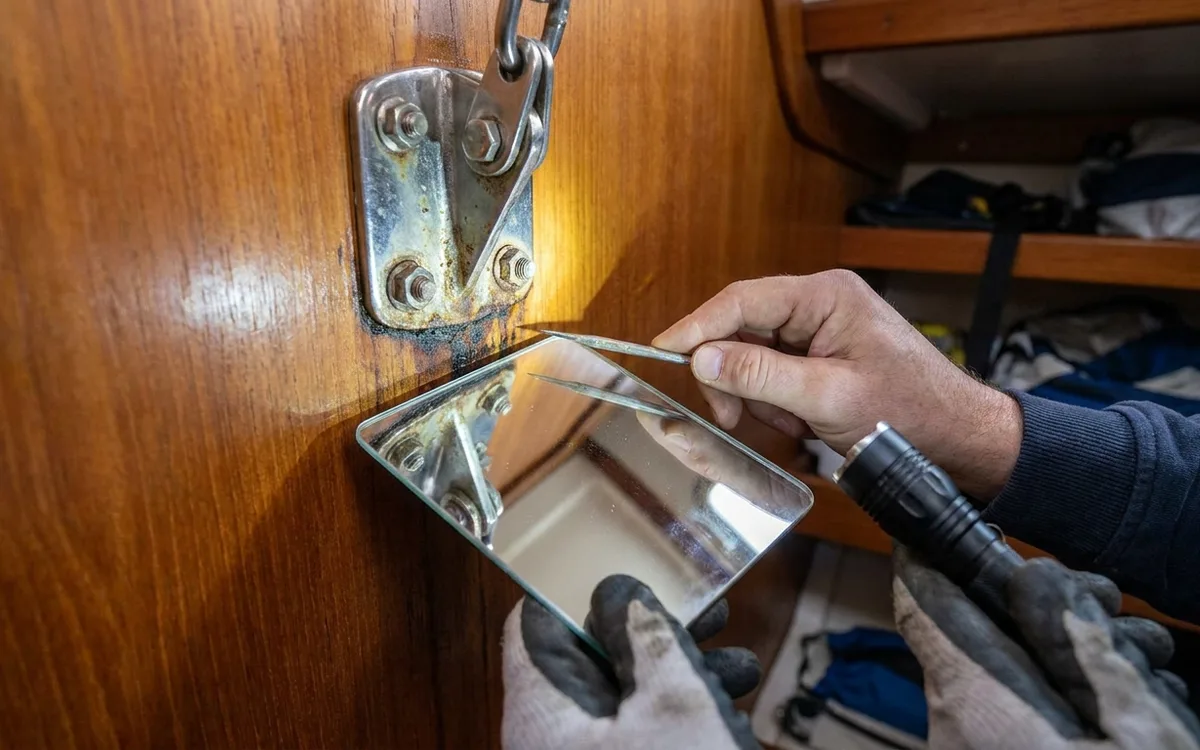 Hands inspect the plywood bulkhead and chainplate bonding inside an opened cabin locker.