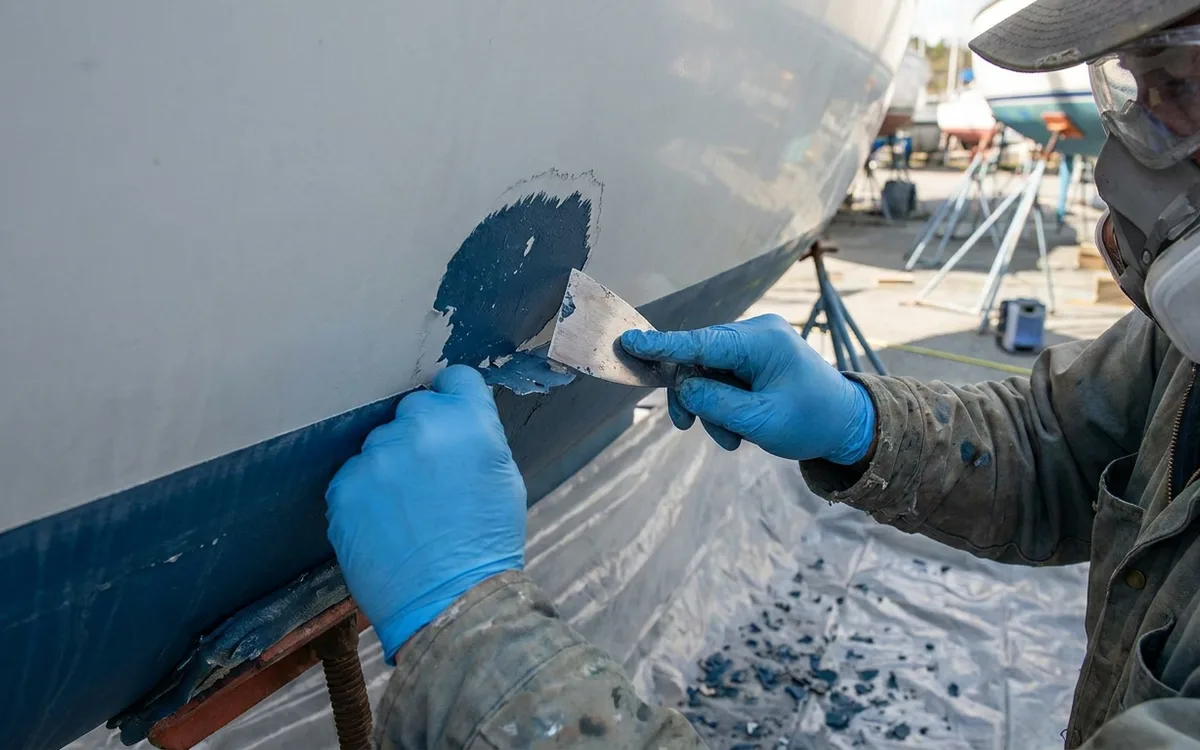 Gloved hands scrape loose antifouling to a firmly attached edge over a debris-catching sheet.