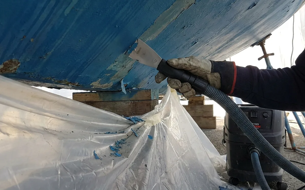Gloved hands use a vacuum-connected carbide scraper nearly flat to remove loose antifouling over secured sheeting.