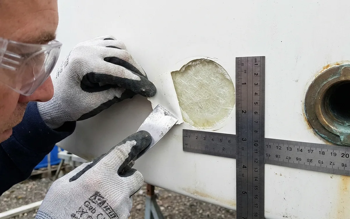 Gloved hands inspect a shallow gelcoat blister beside a ruler and through-hull fitting.