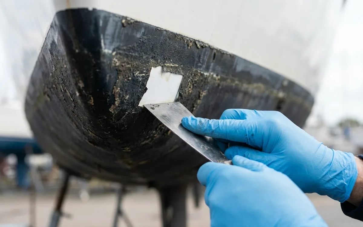 A gloved hand tests antifouling adhesion with a scraper on a small hull patch.