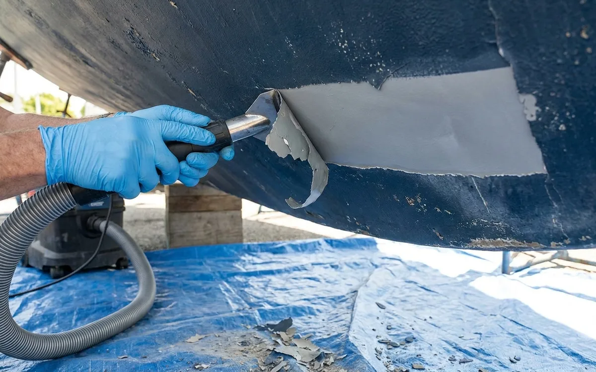 Gloved hands use a vacuum scraper flat to the hull, removing loose antifouling while leaving sound primer intact.