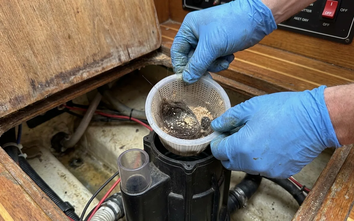 Hands remove hair and sawdust from a bilge pump strainer with the power isolated.