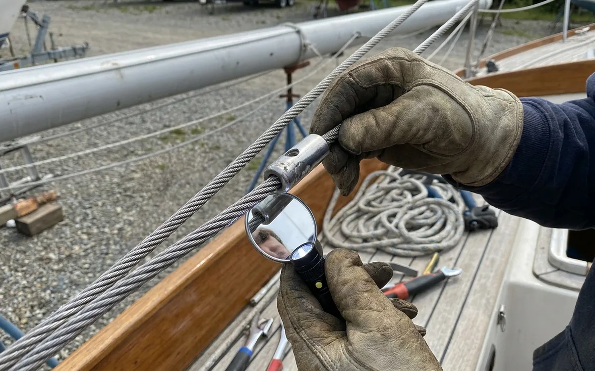 A rigger uses a mirror and flashlight to inspect where a stainless shroud enters its swaged terminal.